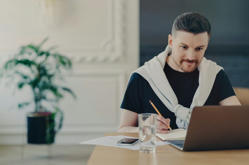 Male office worker looking at laptop and making notes while sitting in office